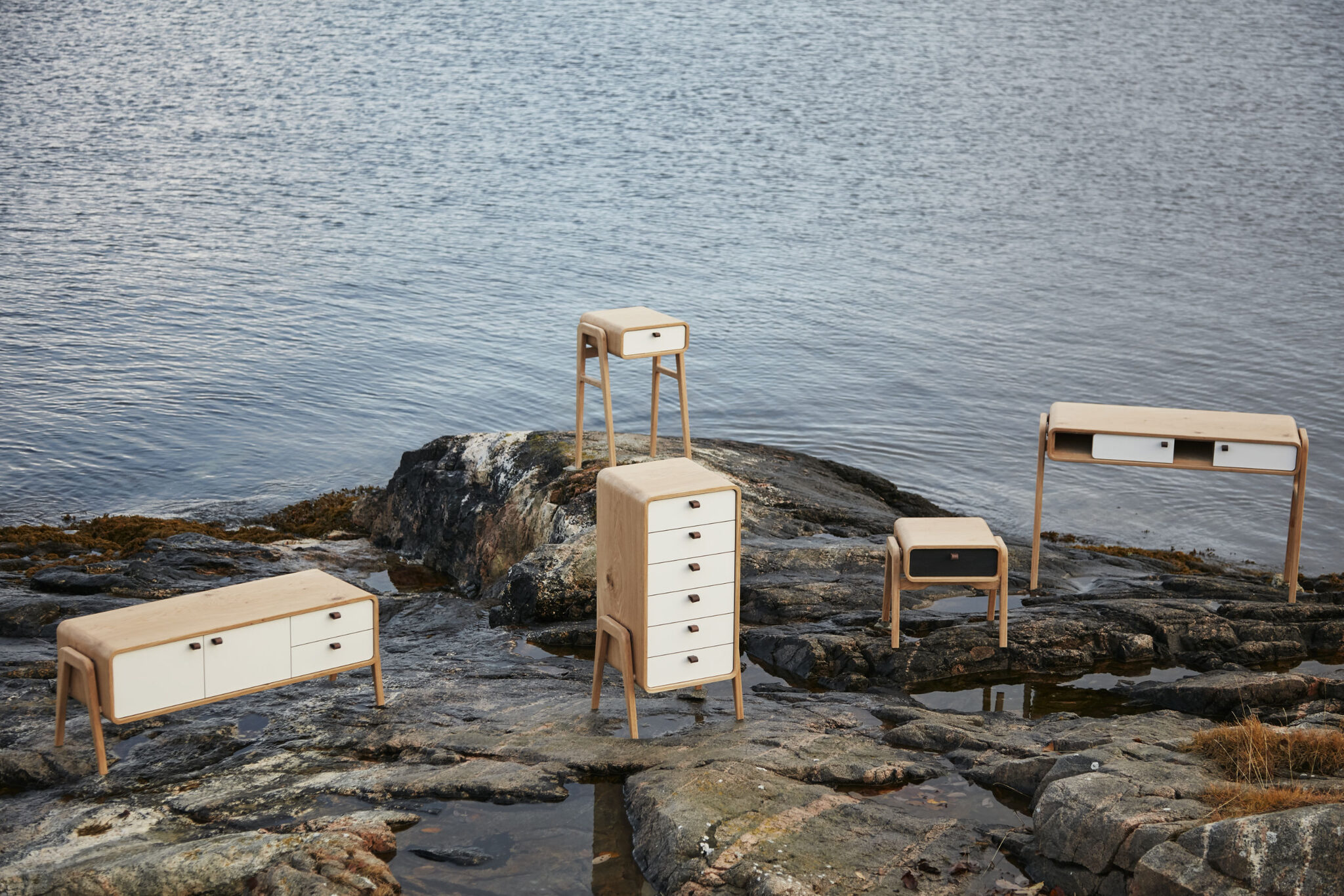 Five distinct furniture models showcased on a coastal rock at the edge of the sea, with water puddles left by the recent tide. nordlys møbler: HANDMADE - SCANDINAVIAN - FURNITURE from Norwegian oak