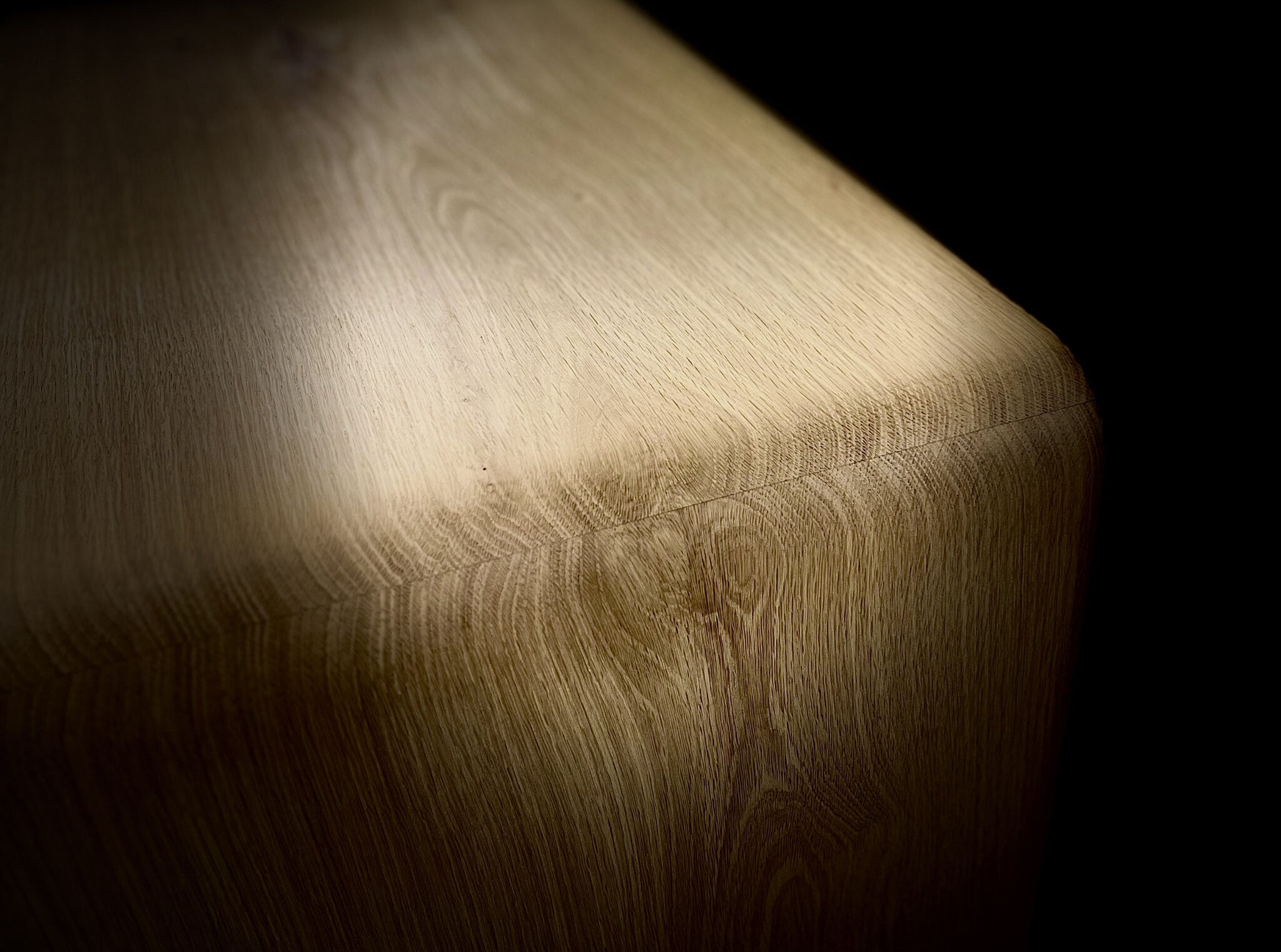 Close-up of a seamlessly rounded oak furniture corner against a dark background, showcasing perfectly continuous wood grain. nordlys møbler: HANDMADE - SCANDINAVIAN - FURNITURE from Norwegian oak 