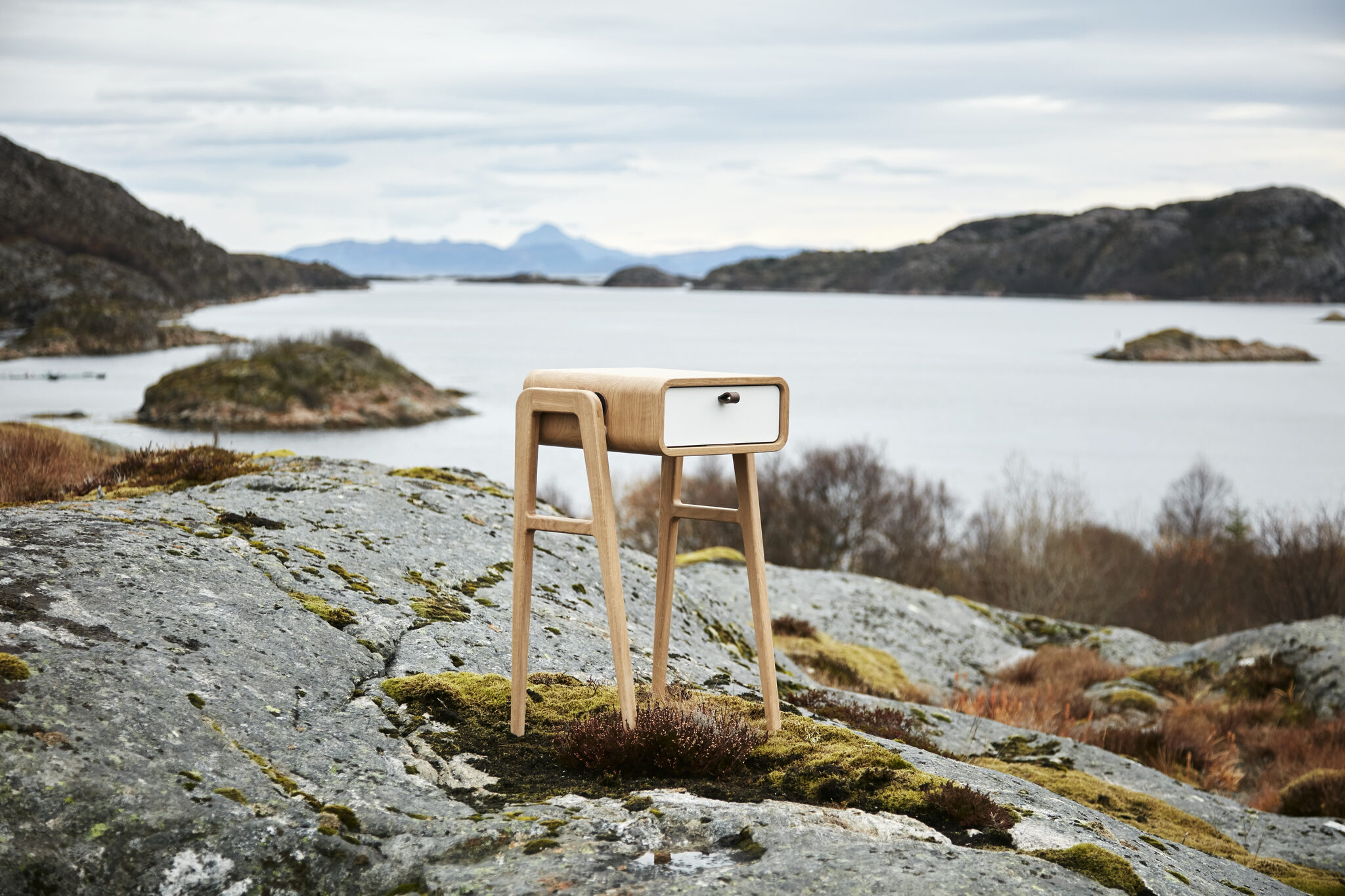White-fronted nm11H nightstand on a mossy rock over a fjord with skerries and Heilhornet view. nordlys møbler: HANDMADE - SCANDINAVIAN - FURNITURE from Norwegian oak.