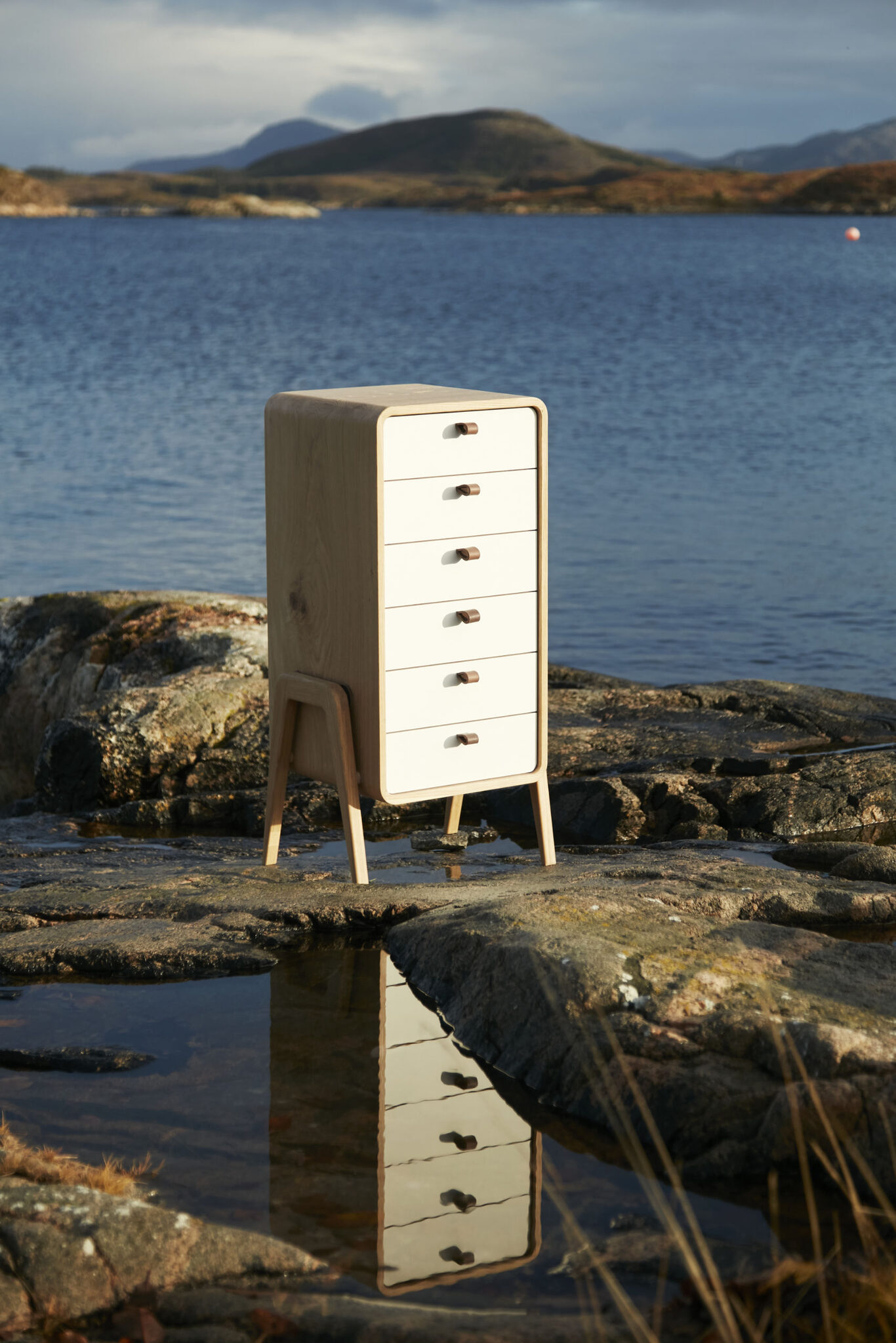nm61L cabinet reflected in a tidal pool against an epic coastal backdrop with mountains. nordlys møbler: HANDMADE - SCANDINAVIAN - FURNITURE, Norwegian oak.